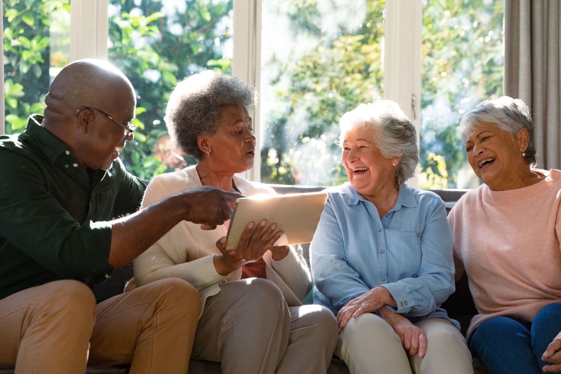 Three happy diverse senior woman and african american male friend sitting on sofa and using tablet