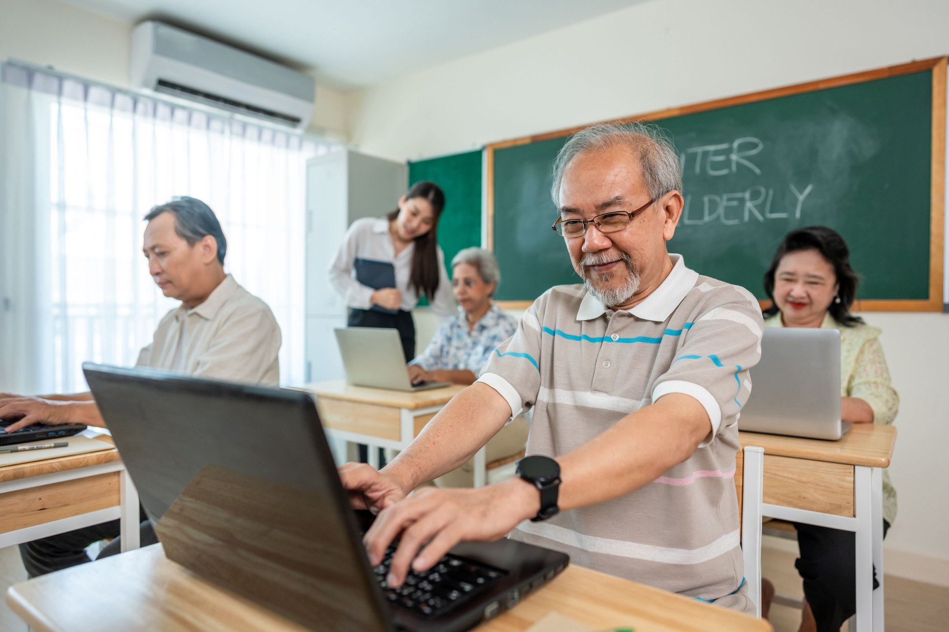 Group of Asian mature students study with instructor in class at school. Senior learners develop new skill and learn to use laptop computer with teacher during adult education course in tutor class.